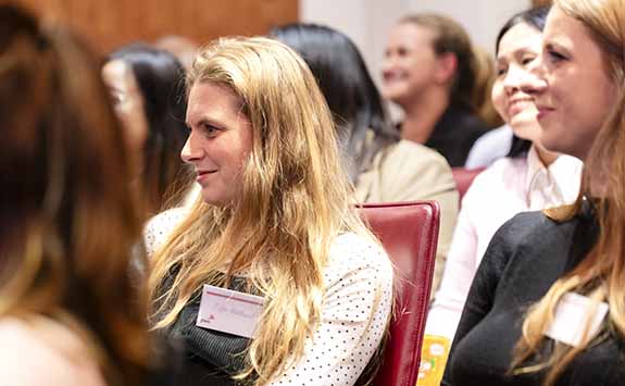 A member of the london alumni network at an event, sitting in a row of seats and smiling towards speaker (off camera)